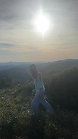 A person stands on a grassy, rocky hilltop in front of a hazy, distant landscape with the sun directly behind them. The person is wearing a light blue, off-the-shoulder top and light blue pants. The person turns their head slightly to the left, and sunglasses are visible. The background is an out-of-focus landscape of hills and greenery under a bright sky.