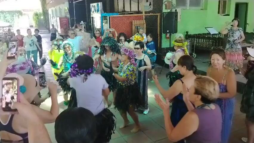 This video captures a lively group of women, most of whom appear to be middle-aged or older, dancing and enjoying themselves at what seems to be an outdoor or semi-outdoor gathering. The setting resembles a patio or covered area, with greenery visible in the background and a tiled floor. Many of the women are dressed in colorful, festive attire, with some wearing floral leis and elaborate headpieces reminiscent of Hawaiian or luau themes. Several individuals are holding up smartphones, seemingly recording the event. The atmosphere is one of celebration and fun, with visible energy and movement from the participants. The lighting suggests it is daytime.