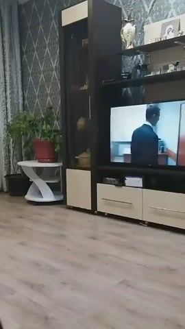A dimly lit room with patterned wallpaper and wood flooring. A large, dark and beige entertainment unit dominates the space, featuring a television that displays a scene of two individuals in what appears to be an office setting. A man in a suit is seen walking towards a door, while a woman is seated at a desk in the background. The entertainment unit is adorned with various decorative items on its shelves, including a vase and picture frames. To the left of the unit, a tall potted plant is visible, and next to it, a small, white, curved table. To the right of the entertainment unit, a small green stool and a low bench are placed against the wall. The camera pans slowly across the scene.