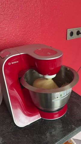 A red and silver Bosch kitchen mixer is actively kneading a ball of dough inside a stainless steel mixing bowl. The mixer is placed on a black countertop against a bright red wall with a double electrical socket. The device is centered in the frame, showing the mixer operating and agitating the dough.