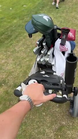 The video shows a person pushing a golf cart along a grassy golf course. The cart is filled with golf clubs and accessories. Other golfers are visible walking in the distance pushing their own carts. The background shows a scenic landscape with trees and hills under a cloudy sky.
