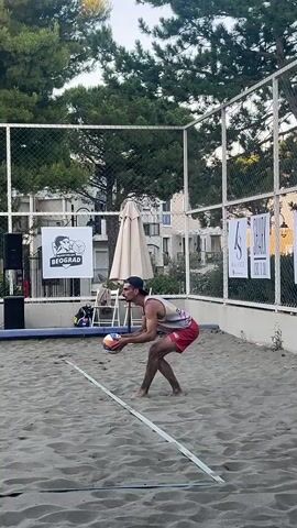 A man is playing beach volleyball. He is wearing red shorts and a sleeveless shirt. He is in a low crouch, preparing to hit the ball. He tosses the ball up and jumps to hit it. He is in the air, with the ball above his head. He lands on the sand. Another man is sitting on a bench in the background. The court is surrounded by a fence and trees. There are buildings in the distance. The sky is clear and blue.