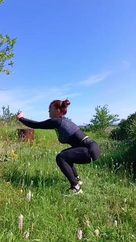 A woman with red hair tied in a bun is performing squats in a grassy field with a clear blue sky. She is wearing a long-sleeved black athletic top with a grey patterned design on the shoulders and chest, and black leggings. Her arms are extended forward for balance during the squats. In the background, there are trees and some wooden structures, one appearing to be an old metal barrel. The scene suggests a sunny day with good weather. The woman appears focused and engaged in her physical activity. The timestamp on the video is visible in the bottom left corner, indicating a duration of a few seconds. The location is identified as Vyazovka, Russia.