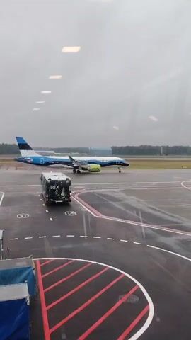A plane with blue, white, and black stripes on its body and tail, and a lime green engine cover, is visible on an airport tarmac on a cloudy day. The plane is parked on the tarmac, with a small white car to the right and a dark grey RIX-branded bus to the left, both also parked. The tarmac is wet, with visible puddles and reflections. The plane is facing right, with its tail towards the left of the frame. The video is shot from inside a building, with reflections of interior lights visible on the window.
