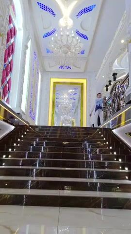 A young man in a blue shirt and white pants walks down a marble staircase. He is on the right side of the frame and is moving towards the camera. The staircase is wide and grand, with ornate railings. The walls are decorated with intricate moldings and illuminated panels. The ceiling features a large chandelier and decorative lighting. The man is holding a phone in his left hand and looking at it. There are other people visible in the background, further down the stairs. The overall impression is one of a luxurious and opulent interior.