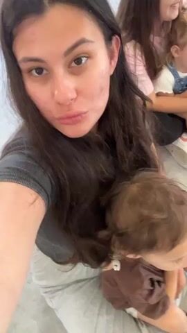 A selfie-style video begins with a young woman holding a baby on her lap. The woman has long dark hair and is wearing a dark grey t-shirt and grey pants. Her face is visible, and she has visible acne on her cheeks and forehead. She is looking at the camera with her lips pursed. The baby has brown hair and is wearing a brown shirt. In the background, another young woman with long dark hair is visible wearing a pink t-shirt, and a toddler wearing blue overalls and a white shirt is partially visible. The environment appears to be indoors, possibly a public space, with a light-colored floor and a white wall in the background. The woman shifts her gaze slightly to the right and then back to the camera, her expression changing from a pout to a slight smile. The video continues to hold on this scene.