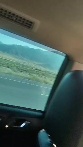 The video shows a view from inside a vehicle looking out the window. The window has a tinted lower portion and an untinted upper portion. Through the window, a road is visible with a white line marking the lanes. Beyond the road, there is a landscape with dry, grassy fields and rolling hills or mountains in the background under a clear blue sky. The sun appears to be shining, casting some light and reflections on the window. The interior of the vehicle is partially visible at the bottom and top of the frame, showing part of a door panel with a handle and a vent. The perspective suggests the vehicle is moving.