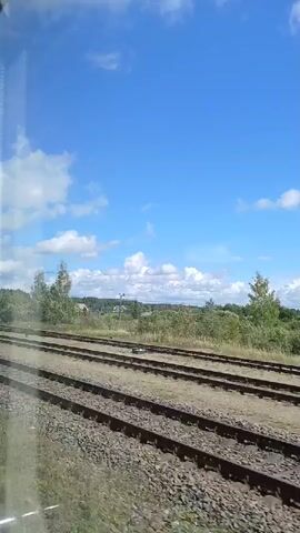 The video shows a view from a moving train. In the foreground, there are multiple sets of train tracks. Beyond the tracks is a landscape with green trees and some buildings in the distance. A tall, metallic tower, possibly a cell tower or radio mast, is visible on the right side of the frame. The sky is blue with some white clouds, indicating a clear day. The camera appears to be positioned looking out of a window on the side of the train.