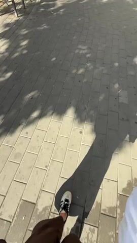 A first-person perspective video opens focusing on a person's feet walking along a paved sidewalk with dappled sunlight. The camera then pivots upwards, revealing a young woman wearing sunglasses, a white short-sleeved henley shirt, and a brown crossbody bag. She is walking on a sidewalk in a sunny urban setting. Behind her are buildings, trees, benches, and a small business front. The camera remains oriented on the woman as she continues walking.