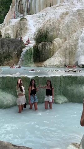 The video shows a view of the natural hot springs of Saturnia, Italy. In the foreground, three young women stand in the milky blue water, posing for a photo. Behind them, more people are relaxing in the pools, some in the cascading water from the natural travertine rock formations. The rock formations are white and creamy, with streaks of green algae, and are covered in lush green vegetation at the top. The water is a pale blue-white color, and steam is visible rising from it. The overall scene depicts a popular tourist destination with a focus on relaxation and natural beauty.