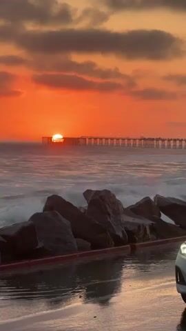 A vertical video opens on a sunset over the ocean. The sun is a bright orange orb near the horizon, partially obscured by a long pier. The sky is filled with dramatic orange and pink clouds. The ocean is wavy, with white foam cresting the waves. In the foreground, large rocks are piled up on the shore. A white car is parked on a wet, paved surface in front of the rocks, with its headlights on. The camera pans slowly to the right, revealing more of the car and the beach. A wave crashes onto the shore, sending spray into the air. The video ends with a view of the car from the side, with the sunset and pier in the background.