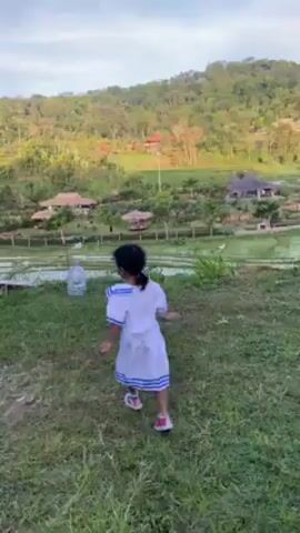A young girl in a sailor dress runs on a grassy hill, stops, turns to the camera, and then runs away. Behind her, there's a terraced rice paddy, various buildings, and a forested hill. A large water bottle sits on the grass near the girl. The sky is blue with some wispy clouds.