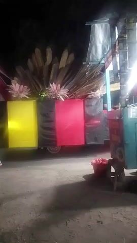 The video shows a night scene with a decorated vehicle. The vehicle is adorned with large, rectangular panels in yellow, black, and red, arranged in a vertical orientation. Above these panels, there is a decorative arrangement of tall, dry, leaf-like elements and some pink floral accents. A flag, appearing to be red, white, and red, is draped on the left side of the vehicle. In the foreground, to the left, a table with a large water dispenser and some boxes is visible. To the right, a street vendor stall with a turquoise-colored structure and signage is present. A red bucket filled with items is also on the ground near the stall. The ground is unpaved and appears to be dirt or gravel. The lighting suggests it is nighttime, with some artificial lights illuminating the scene. There are no clear views of any individuals.