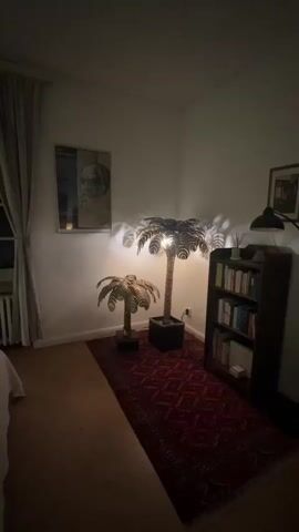 This video shows a dimly lit room with a large window. The window has multiple panes and is partially covered by light-colored curtains. Outside the window, it appears to be nighttime, and a bright moon is visible in the sky. The room contains a bed on the left side, a radiator beneath the window, and a bookcase filled with books on the right side. Two palm tree-shaped lamps are positioned in front of the bookcase, casting a warm glow. A framed picture of a man's face is hanging on the wall above the lamps. A red patterned rug covers a portion of the floor in front of the bookcase and lamps. The overall ambiance of the room is cozy and serene, with the moonlight adding a touch of magic to the scene.