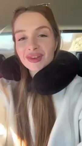 A woman with long hair is wearing a black travel pillow. She has red nail polish and is wearing a white sweatshirt. She's talking to the camera and is inside a vehicle. The interior of the vehicle is visible in the background.
