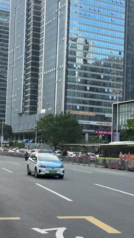 The video shows an urban street scene. Tall buildings dominate the background, their glass facades reflecting the sky. A white and blue electric taxi drives past on the road. A yellow bus is also visible, stopped along the side of the road, and several cars are parked nearby. The road has white lane markings and yellow directional arrows. A tree provides some greenery amid the urban landscape. The scene appears to be taken during the daytime under bright sunlight.