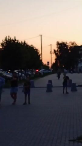 A slightly out-of-focus video shows a street scene at sunset. The sky is a warm gradient of orange and pink. Silhouetted trees line the left side of the street, with parked cars visible behind them. A paved walkway runs down the center of the frame, with several cylindrical, concrete bollards placed at intervals. Several figures, appearing to be people, are scattered along the walkway. On the left side, two figures, one taller and one shorter, stand near each other. Further down the walkway, a single figure is visible. On the right side of the frame, a few more figures are present, walking and standing. Buildings with shopfronts are visible on the far right, also in silhouette. The overall impression is a peaceful evening stroll or gathering on a street as the day ends.