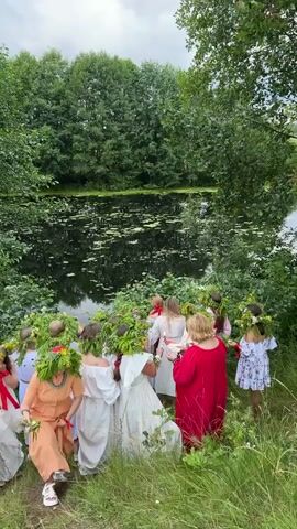 A group of people, mostly women, are gathered outdoors near a body of water surrounded by trees. Many of the individuals are wearing white or light-colored dresses and floral wreaths on their heads. One woman in the foreground wears an orange outfit. Some of the women are holding what appear to be candles. The group seems to be walking in a procession or gathered for an event. The environment is lush and green, suggesting a natural setting.