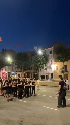 The video captures a group of people dressed in black t-shirts with logos on them, standing in a street at dusk. They appear to be holding sheet music or similar items in their hands, suggesting a performance or rehearsal. One person, also in black, stands facing them, seemingly directing or leading. The street is paved and marked with white lines. Buildings line the street, illuminated by streetlights and interior lights. In the background, there are some decorative elements hanging from lampposts, including what looks like a red floral design and a yellow floral design. The sky is a deep blue, indicating that it is evening. Some members of the group are looking at the person facing them, while others are looking down at their papers. The overall atmosphere seems to be one of anticipation and preparation for an event.