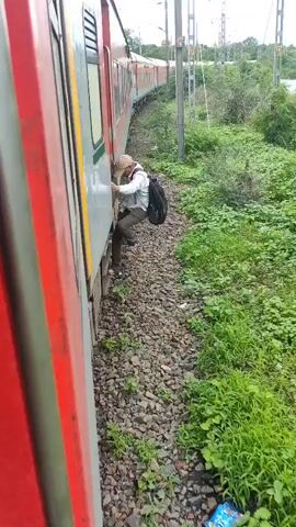 A person is shown clinging to the outside of a moving train. They are wearing a light-colored shirt, dark pants, and a backpack. The train is traveling along a track lined with green vegetation and rocks. The sky appears cloudy. Another person's hand, wearing a watch, is visible near the camera, suggesting the video is taken from inside the train. The person outside the train is facing away from the camera and appears to be struggling to hold on.