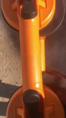 A hand holds an orange, dual-suction cup lifter with black rubber rings. The device has two handles with black levers, and the surface it is resting on appears to be concrete. The lighting is bright.