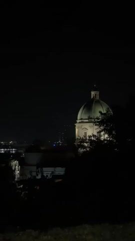 The video shows a dimly lit night scene with a prominent green domed building in the center. The dome is illuminated by artificial lights, casting a warm glow. In the foreground, dark silhouettes of trees and foliage obscure a portion of the view. In the background, distant city lights are visible, suggesting an urban environment. The overall mood is serene and mysterious due to the darkness and the isolated illumination of the building.