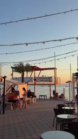 The video shows a beach scene during sunset. In the foreground, there are tables and chairs for an outdoor cafe. A group of people are sitting at a table on the left, with a woman in a white dress standing nearby. In the middle ground, there is a red lifeguard tower with a sign that reads "SPASATEL'NY POST RESCUE POST". Two people are sitting on swings attached to the tower, and another person is standing on the tower platform. Further back, several people are walking on the beach and in the water, with boats visible on the ocean. The sky is clear and a pale orange hue from the sunset. String lights with bulbs are visible overhead.