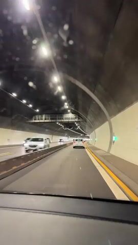 The video shows the perspective from inside a vehicle driving through a tunnel. The tunnel is well-lit with overhead lights and has multiple lanes of traffic. Several cars are visible in front and to the side of the vehicle. The walls of the tunnel are made of concrete and appear to be curved. There are yellow lines marking the lanes and a concrete barrier separating the lanes. The vehicle is moving forward at a moderate speed.