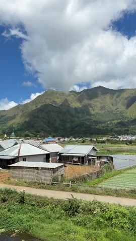 The video shows a rural landscape with houses, fields, and a mountain range in the background. The houses have simple structures and tin roofs. The fields are green and appear to be cultivated. The mountains are covered in lush vegetation, with clouds partially obscuring their peaks. The sky is blue with fluffy white clouds. The camera slowly pans to the right, revealing more of the agricultural land and the mountains. The overall impression is that of a peaceful and scenic countryside.