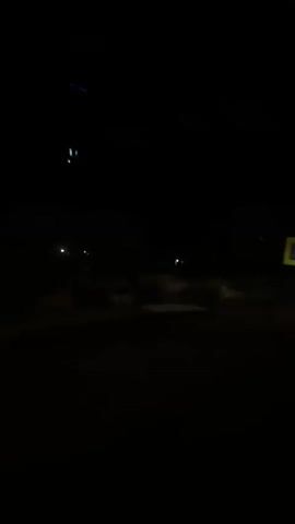 The video is a nighttime recording taken from inside a moving vehicle. The scene outside is dark, with only faint illumination from distant lights. A yellow square traffic sign with a blue symbol is visible on the right side of the frame. The road appears to be unpaved or dirt. The perspective suggests the vehicle is driving past a rural or undeveloped area.