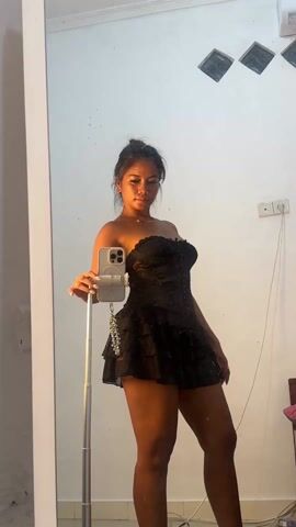 A woman stands in front of a mirror holding a phone on a stand. She poses in a short, strapless black dress with a corset top and ruffled skirt. Her hair is pulled up in a bun. The background shows a white wall with an air conditioner, an electrical outlet, and decorative wall panels. The mirror has a white frame.