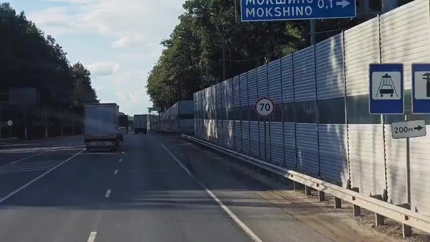 A truck travels on a highway alongside a sound barrier. The sound barrier is made of metal panels and appears to be a significant length of the road. The truck is in the right lane of the highway. The sky is partly cloudy and the sun is visible. There are trees on the left side of the road and on the other side of the sound barrier.