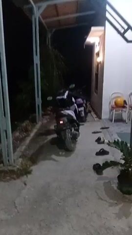 The video shows an outdoor scene at night. Two motorcycles are parked under a covered area. In the background, there is a building with lights on. Two chairs with red cushions are placed in front of the building, with a yellow helmet on one of them. Several pairs of flip-flops are scattered on the ground. The ground is paved with concrete and tile.