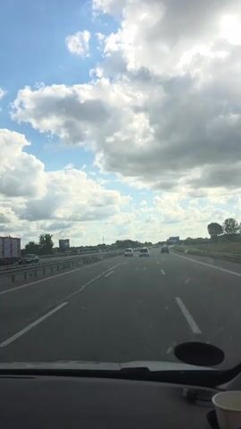 This video captures a first-person perspective from inside a vehicle traveling on a highway. The scene is dominated by a bright, partly cloudy sky with large, fluffy white clouds interspersed with patches of blue. The sun is visible, suggesting it is daytime, likely late morning or afternoon, given the brightness and the long shadows cast by the guardrail on the right side of the road.

The highway itself is paved and appears to be in good condition, with clear lane markings. Several cars are visible on the road ahead, traveling in the same direction. The vehicles are mostly white or light-colored, and their movement suggests moderate traffic flow. A guardrail runs along the left side of the highway, separating it from an adjacent area that appears to be fields or open land. On the right side, trees and vegetation line the roadside, with a distant, tall, rectangular building visible in the far left background, potentially a silo or industrial structure. A road sign is also visible in the distance on the right side of the highway.

The overall mood is one of travel and a pleasant, somewhat dynamic sky. The focus is on the journey itself, with the natural elements of the sky and the man-made structure of the highway and vehicles providing the visual content. There are no people clearly visible outside of the cars, and the interior of the vehicle is only partially seen at the bottom of the frame, showing a dashboard and a white cup. No text is clearly discernible in the video. The provided location context, Skanderborg, Denmark, suggests this highway segment is within that region.