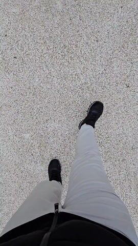 The video begins with a view of someone's legs and shoes standing on a grey-pebbled surface. They are wearing white pants and black shoes. The camera then tilts up to reveal a path leading to a white clock tower. The path is lined with low hedges. On either side of the path are white buildings, which appear to be shops. The sky is overcast. The scene is deserted except for the person recording the video.