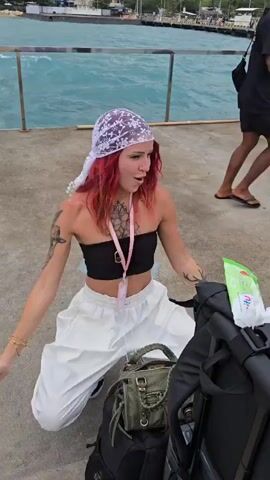 A young woman with red hair kneels on a pier. She has a black strapless top, white baggy pants, and a white lace bandana. She is surrounded by luggage, including a black suitcase on wheels, a black backpack, and a smaller green bag. She is smiling and appears to be reaching out to someone passing by. A man in a black t-shirt and patterned shorts walks behind her. The pier has a metal railing, and the water is visible in the background. There are buildings visible in the distance.