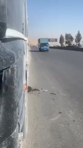 The video shows a roadside view, presumably from a parked or slowly moving bus. The camera focuses along the side of the bus, partially visible on the left, while a highway stretches to the right. Other vehicles, including a blue truck and several cars, are visible on the road, and a sign is seen in the distance. The environment appears to be a dry, possibly desert-like area, with a hazy sky.