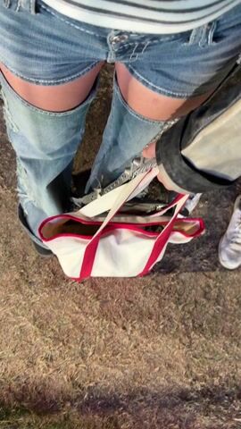 The video shows a person wearing ripped jean shorts and a striped shirt standing outdoors. They are holding a white tote bag with red handles. The ground is covered in dry grass. A black jacket is draped over the tote bag. The person also appears to be wearing sneakers.