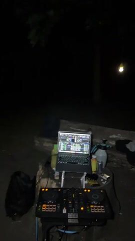 A dj setup is visible outdoors at night. The setup includes a laptop, a dj controller, and various other electronic devices and accessories. The lighting is dim, with a single bulb hanging in the background. The ground appears to be dirt or gravel, and there are trees and foliage visible in the background. There are no people visible in the scene. The dj controller is a Pioneer DDJ-SX3, and the laptop is a Macbook Pro. The scene is static, with no movement or action captured.