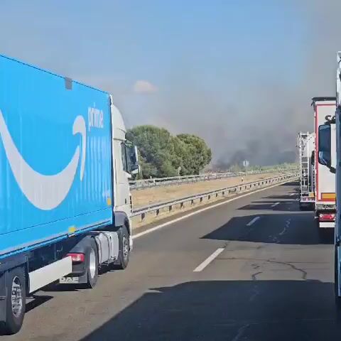 A white truck with the Amazon Prime logo is driving on a highway. The truck is in the foreground on the left side of the frame. In the background, there is a large plume of smoke rising from a fire. The fire appears to be in a dry, grassy area with some trees. The sky is hazy and overcast with smoke. There are also guardrails along the side of the highway, and a few traffic signs are visible. The overall scene suggests a wildfire event occurring near a roadway.