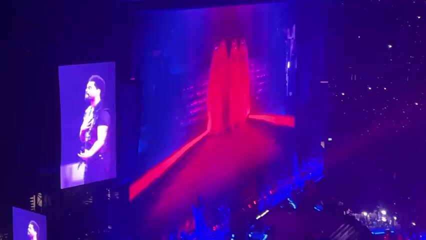 A concert stage is illuminated with bright red and blue lights. A large screen on the left displays a performer in a black outfit, his hand on his chest. The main screen shows a performer in a silver top and black pants, also with a hand on his chest, standing near three figures in flowing red robes. The performer then turns and walks away from the figures. The stage is adorned with red lights creating a pathway.