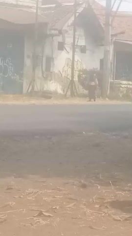 A hazy video shows a street scene. On the left, a building with a blue garage door covered in graffiti stands next to a smaller building. In front of the buildings, a few people are visible. One person is riding a scooter, wearing a white helmet and a dark outfit. Further down the street, another person in a light-colored outfit is walking. On the right side of the street, a white car is parked. The ground in the foreground is dirt and gravel.