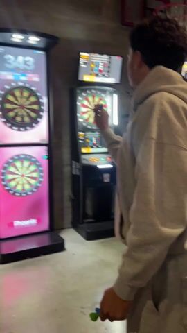 A person with dark curly hair, wearing a light-colored hoodie, is playing darts at an electronic dartboard indoors. The individual is positioned to the right of the frame, facing the dartboard, and is in the process of throwing a dart.

There are two electronic dartboards visible; the one on the left is the primary focus. Its top screen displays game information for a "501 GAME" in "ROUND 4 / 15". Player scores are shown as Player 1 with 350 points, Player 2 (the person playing) with 340 points, and Player 3 with 398 points. The screen updates to show a "TRIPLE 1" hit, scoring 3 points, followed by a "20" point hit. The bottom screen of this dartboard unit clearly displays "VS Phoenix X". Another similar electronic dartboard is visible to the right and slightly further back. A partial view of a basketball hoop or backboard is visible in the upper right background. The walls appear to be concrete or a similar material, and the floor is concrete, suggesting an indoor recreational or entertainment venue.
