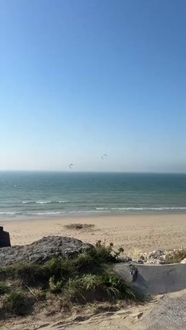 This video shows a serene beach scene with the ocean as the primary focus. The sky is a clear, bright blue, suggesting a sunny day with good weather. The sea is a deep blue-green, with gentle waves lapping at the sandy shore. There are several kite surfers in the water, their colorful kites forming arcs against the horizon. They appear to be enjoying the wind and the waves, engaging in the sport of kitesurfing. The beach itself is sandy and appears mostly empty, adding to the sense of peace and tranquility. In the foreground, there are some rocks and vegetation, hinting at the natural beauty of the location. The overall impression is one of a peaceful and enjoyable day at the beach, with the activity of kitesurfing adding a dynamic element to the otherwise calm scene. The video captures the essence of a perfect day for water sports and relaxation. The time of day appears to be daytime, likely mid-morning to early afternoon, given the bright sunlight and clear skies. The location context places this scene in Wissant, France, a popular spot for kite surfing.