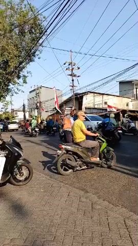 A street scene shows several motorbikes and a car on a paved road. Overhead power lines crisscross the sky. Two individuals are visible in the foreground, one on a green motorbike and another on a black motorbike. In the background, several more motorbikes are moving in traffic. Two people appear to be holding Indonesian flags. The overall impression is of a busy street with people celebrating or participating in an event.