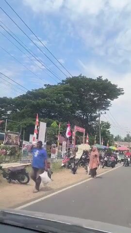 The video shows a street scene with a large tree in the background. There are several motorcycles parked along the side of the road, and a few people are seen walking and standing near stalls set up under umbrellas. Indonesian flags are displayed, suggesting a national celebration or event. The sky is partly cloudy. The camera appears to be inside a vehicle, as evidenced by the dashboard visible at the bottom of the frame.