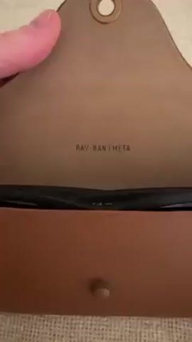 The video shows a person closing a Ray-Ban Meta sunglasses case. The case is brown leather. The inside of the case has the words "RAY-BAN | META" printed on it. A green light turns on when the case is closed. The case is sitting on a textured tan surface. A person's hand closes the case.