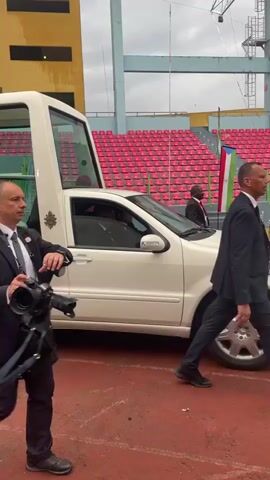The video captures an outdoor scene at what appears to be a stadium or large arena in Malabo, Equatorial Guinea. A white customized vehicle, resembling a Popemobile with a tall, clear glass enclosure at the rear, is central to the frame. Inside the glass enclosure, a man dressed in white robes, likely the Pope, is seated and visible to the public.

Several men in dark suits are walking alongside and near the vehicle. In the foreground, a man in a dark suit with an earpiece is walking towards the right, holding a professional camera with a large lens, suggesting he is a photographer or media personnel. Another man in a suit is walking closely beside the rear passenger side of the customized vehicle. A third man in a suit is visible walking further behind the vehicle.

The background features rows of red and blue stadium seating, a red track or pavement surface, and parts of a larger structural framework. The sky is overcast, indicating a daytime event under cloudy conditions. The overall atmosphere appears to be one of security and official protocol surrounding a public appearance. The flag of Equatorial Guinea is faintly visible in the distance among the stadium seats.