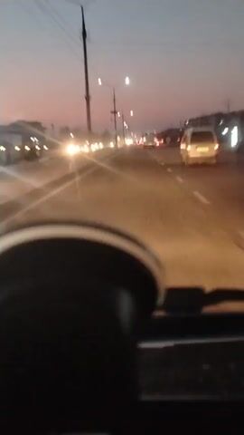 The video shows a drive at dusk. It is a driver's point of view of the road in front.  Other cars are visible, some with headlights on.  Streetlights are visible on either side of the road. The dashboard and steering wheel are visible in the foreground.