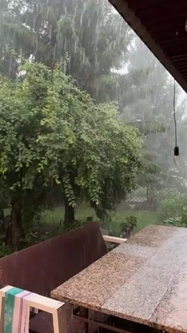 A heavy rain is falling outdoors, obscuring the view of trees and foliage in the background. In the foreground, a granite-topped table sits under a covered porch. The rain is visible as streaks falling from the sky.